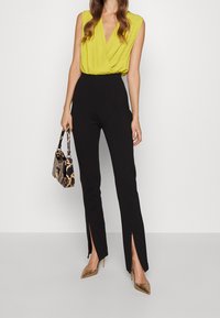 Haut sans manches jaune avec un design cache-cœur, associé à un pantalon noir taille haute avec des fentes latérales, des escarpins dorés pointus et un sac à main à motif.