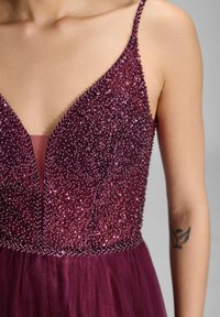 Cekinowa burgundowa sukienka z głębokim dekoltem w kształcie V, cienkimi ramiączkami i teksturowanym górnym odcieniem. Lekką, przezroczystą spódnicą z delikatnym blaskiem i haftowanymi detalami.
