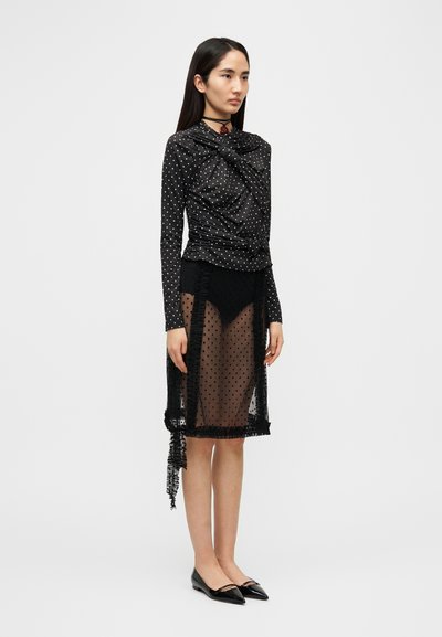 Μαύρο πουκάμισο με λευκά πουά και σχεδιασμό με ραμμένο ύφασμα, συνδυασμένο με διάφανο μαύρο φούστα με κρόσια στο τελείωμα. Μαύρα παν Flats ολοκληρώνουν την εμφάνιση.