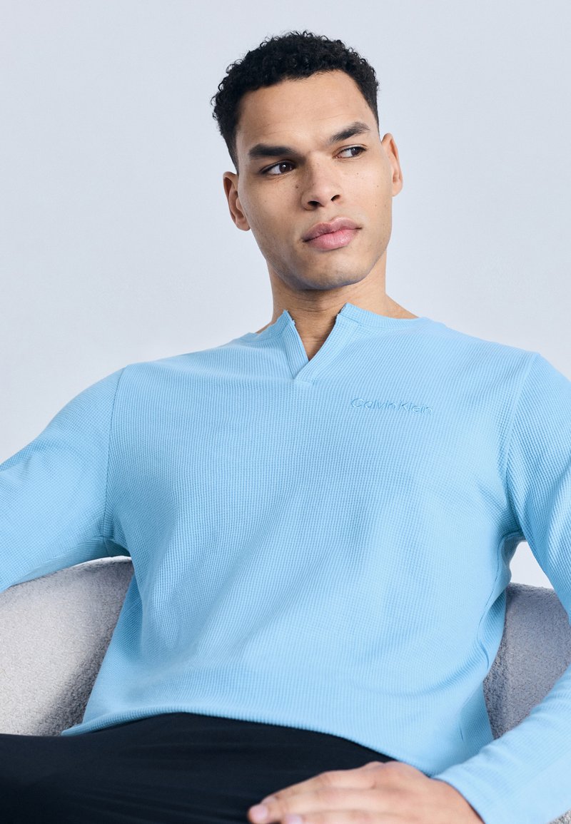 Άνδρας με κοντά, σγουρά μαλλιά καθισμένος, φορώντας ανοιχτό μπλε πουκάμισο Calvin Klein μ ε μακριά μανίκια, κοιτάζοντας προς τα πλάγια μπροστά σε ανοιχτό γαλάζιο φόντο.