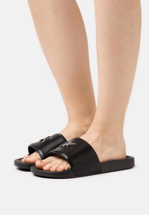 Pieds portant des sandales slides noires avec une large lanière et un logo métallique embossé sur un fond blanc uni.