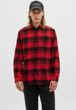 Joven con cabello largo que lleva una gorra negra, una camisa de cuadros rojos y negros abotonada, pantalones negros y anillos y tatuajes en las manos.