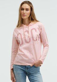 Hellrosa Kapuzenpullover mit langen Ärmeln, großem Text und Kordelzug am Kapuzenbereich. Weiches Material mit legerer Passform und dezentem Logodetail.