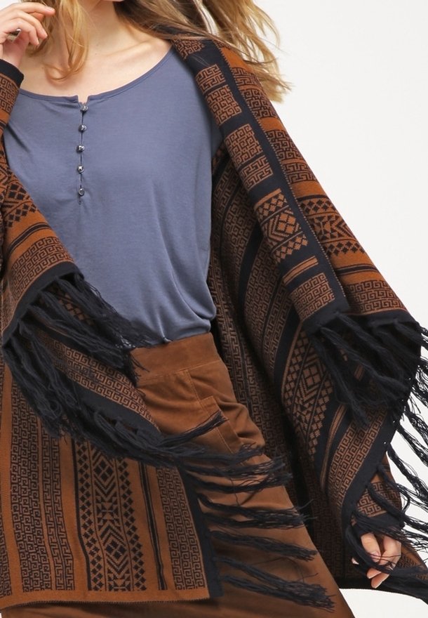 Femme portant un châle marron à motifs avec des franges noires sur un haut bleu boutonné et une jupe marron, tenant le châle ouvert.