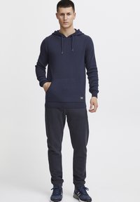 Sudadera azul marino de tela suave con un bolsillo frontal, cordones y puños acanalados. Combinada con pantalones de chándal azul marino y zapatillas deportivas oscuras.