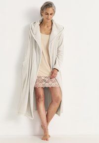 Ljusgrå fleecekappa med en stor huva, sidfickor och en avslappnad passform, bärs över en beige klänning med en spetsdekoration.