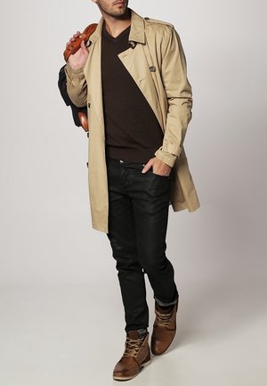 Beige trenchcoat over een donkerbruine trui, gecombineerd met zwarte spijkerbroeken en bruine laarzen. De jas heeft dubbele knopen en een uitlopende kraag.