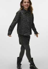 Une jeune fille souriante marche dans une veste et une jupe à imprimé léopard noir, des collants gris foncé et des bottes noires à boucle, sur fond blanc.