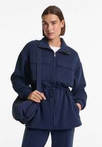 Veste zippée bleu marine avec deux poches poitrine, taille élastique et manches longues. Associée à un sac assorti en tissu similaire.