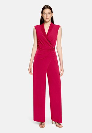 Kobieta ubraną bezrękawny głęboko różowy kombinezon z szerokimi nogawkami i wiązanym detalem w talii, zestawiony ze złotymi sandałami na wysokim obcasie z paskami.