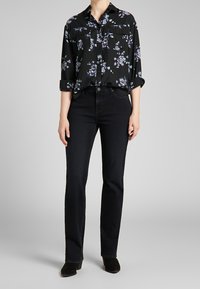 Svart blommig blus med lila accenter, två bröstfickor, knäppning framtill, kombinerad med mörka jeans och svarta mockaskor.