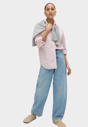 Mujer con una camisa de rayas rosas, vaqueros azules de pierna ancha, zapatos beige sin cordones y un suéter gris sobre los hombros.