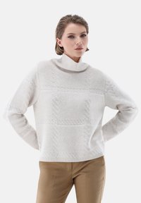 Cremfarbener Rollkragenpullover mit strukturiertem Strickmuster und geripptem Saum. Kombiniert mit beigen, taillierten Hosen, die eine entspannte Passform zeigen.