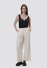 Top senza maniche nero con design floreale; pantaloni larghi crema; sandali neri spessi. Modella in piedi con le mani in tasca su uno sfondo neutro.