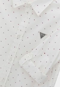 Biała koszula z guzikami, ozdobiona małym czerwono-niebieskim geometrycznym wzorem, z złożonym mankietem i trójkątną naszywką z logo po lewej stronie klatki piersiowej.