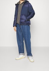 Granatowa pikowana kurtka z teksturowanym wykończeniem, wyposażona w kaptur, zapinana na zamek z przodu, z dwoma bocznymi kieszeniami, noszona na szarej bluzie z kapturem i luźnych niebieskich dżinsach.