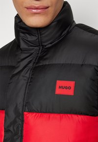 Gros plan sur une personne portant une doudoune noire et rouge avec un patch logo "HUGO" rouge sur la poitrine.