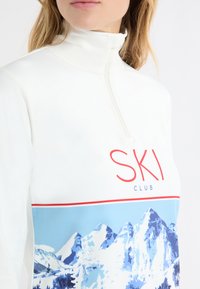 Biały pulower na pół zamek z grafiką "SKI CLUB", przedstawiającą niebieski górski krajobraz na dolnej części oraz miękką, gładką fakturą.