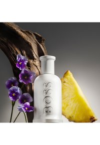 Bouteille BOSS blanche, fleurs violettes, bois flotté incurvé et une tranche d'ananas jaune frais sur un fond dégradé gris.