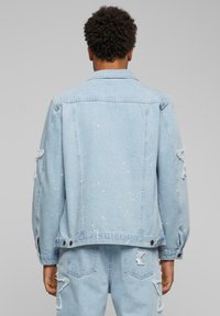 Light blue denim jacket featuring distressed details, scattered white paint splatters, and button closures at the cuffs and hem.