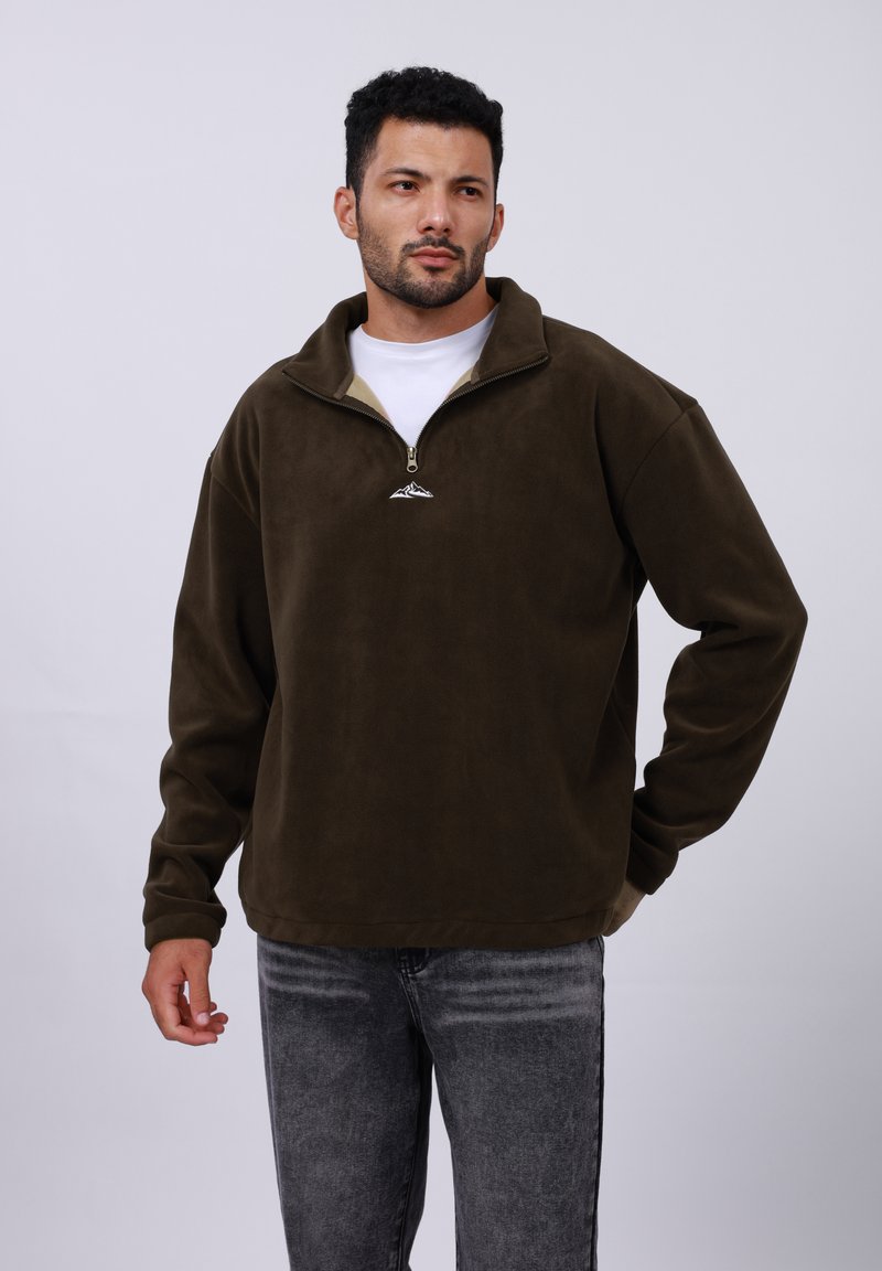 Pullover en polaire zippé marron avec un col haut, présentant une doublure beige clair et un petit logo de montagne sur la poitrine. Jeans gris.