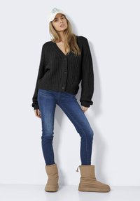 Zwarte gebreide cardigan met een v-hals en knopen, gecombineerd met een blauwe skinny jeans en tan enkellaarsjes. De algehele uitstraling is casual en warm.