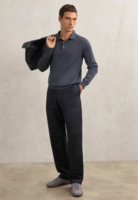 Tumši pelēks polo krekls ar garām piedurknēm, tumši zili platām kājām bikses un pelēki sporta apavi. Modelis tur melnu jaku pāri plecam, fonā ir fakturēta aizmugure.