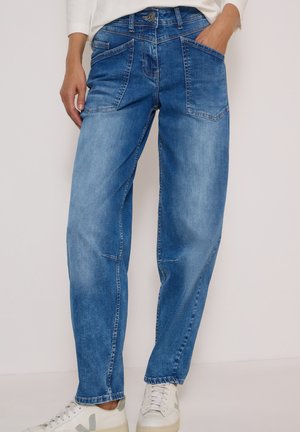 Jean en denim bleu avec poches plaquées devant, porté par une personne avec une main dans la poche et des baskets blanches aux pieds.