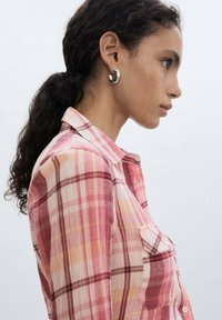 Pink and orange plaid shirt with a collar, featuring long sleeves and a chest pocket. Model has curly hair and wears silver hoop earrings.