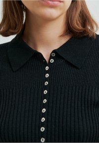 Svart rebbad cardigan med krage och silverknappar fram. Texturen är stickad, med vertikala ribbningar och eleganta detaljer.