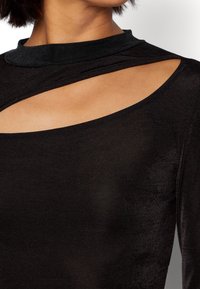 Svart långärmad topp med hög krage, med en utskärning på bröstet. Tillverkad av mjuk, lättviktsmaterial med en subtil lyster.