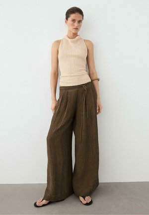 Donna in piedi contro un muro bianco, indossa un top beige in maglia senza maniche, pantaloni larghi marroni con texture, infradito nere e un braccialetto di legno.
