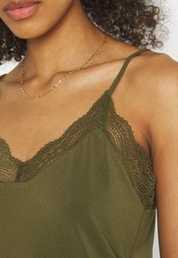 Femme portant un débardeur en dentelle olive et un délicat collier en chaîne dorée, avec des cheveux bouclés visibles sur un fond clair.