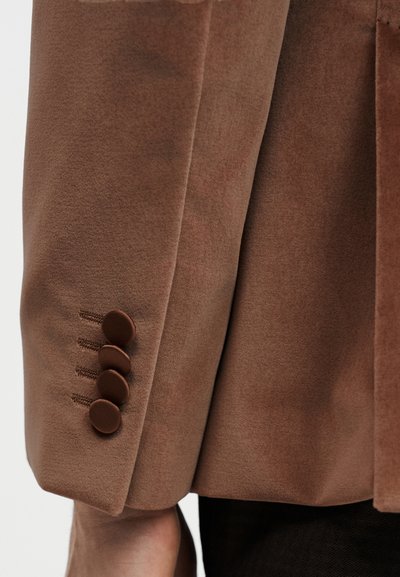 Μανίκι καφέ σακακιού που αναδεικνύει την απαλή υφή του υφάσματος, με τέσσερα κουμπιά καλυμμένα με ύφασμα και διακοσμητική ραφή.