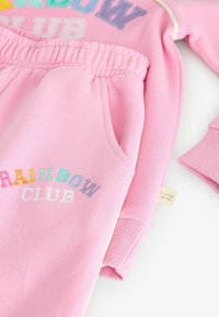 Šviesiai rožinis sportinis kostiumas su elastinga juosta, šoninėmis kišenėmis ir įvairiaspalve "RAINBOW CLUB" siuvinėjimu. Minkšta, struktūrinė medžiaga.