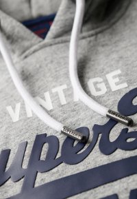 Felpa grigia con cappuccio e lacci bianchi, caratterizzata da scritte blu scuro. I lacci hanno estremità in metallo incise con "SUPERDRY." Texture morbida.