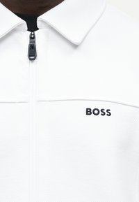 Άσπρο ένδυμα με φερμουάρ και γιακά, με κεντητό μαύρο λογότυπο BOSS στο αριστερό στήθος και μαύρο τσέπωμα φερμουάρ.