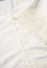 Tissu blanc à boutons avec un détail en dentelle texturée présentant des motifs ovales et circulaires sur le côté.