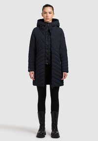 Gequiltede donkerblauwe lange jas met capuchon, voorzien van chevronpatronen, een rits aan de voorkant en zijzakken. Gestyled met zwarte leggings en laarzen.