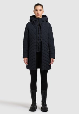 Gequiltede donkerblauwe lange jas met capuchon, voorzien van chevronpatronen, een rits aan de voorkant en zijzakken. Gestyled met zwarte leggings en laarzen.