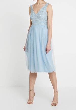 Femme portant une robe bleu clair sans manches, longueur genou, avec un corsage orné de perles et une superposition en tulle, associée à des escarpins nus à bride autour de la cheville.