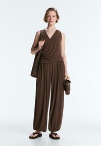 Femme debout portant un haut marron sans manches à nouer, pantalon ample, sandales compensées, tenant un tapis de yoga sur l'épaule et une bouteille d'eau.