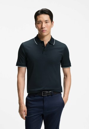 Mand iført en sort poloshirt med hvid kant og marineblå bukser, stående med den ene hånd i lommen mod en ensfarvet hvid baggrund.