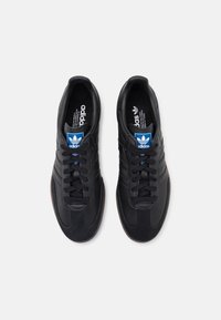 Black leather sneakers with a textured black suede toe, featuring three stripes, blue tongue label, and brown rubber outsole.