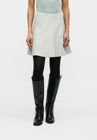 Γυναίκα που φοράει ανοιχτό γκρι φούστα σε γραμμή Α, μαύρες αδιαφανείς καλσόν και μαύρες δερμάτινες μπότες μέχρι το γόνατο, σε απλό φόντο.