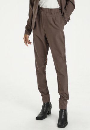 Personne portant un pantalon marron à taille élastique, une veste marron, un haut blanc et des bottines à talons noires, debout devant un fond uni.