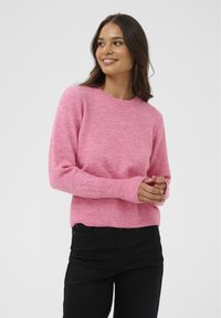 Gaiši rozā džemperis ar apaļu kakla izgriezumu, garām piedurknēm, ribotām aprocēm un apakšmalu, ar mīkstu teksturētu adījumu. Valkāts ar melnām biksēm.