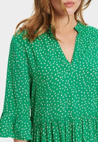 Zelené šaty z lehkého materiálu s bílými puntíky. Mají véčkový výstřih, volánové rukávy a sukni s vrstveným designem.