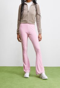 Person som bär en beige zip-up jacka, vita sneakers och högmidjade rosa utsvängda byxor, står på en grön matta mot en vit vägg.