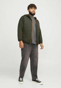 Veste matelassée vert foncé avec fermeture éclair orange, associée à une chemise grise et un jean noir. La veste a une capuche et des poignets élastiques.